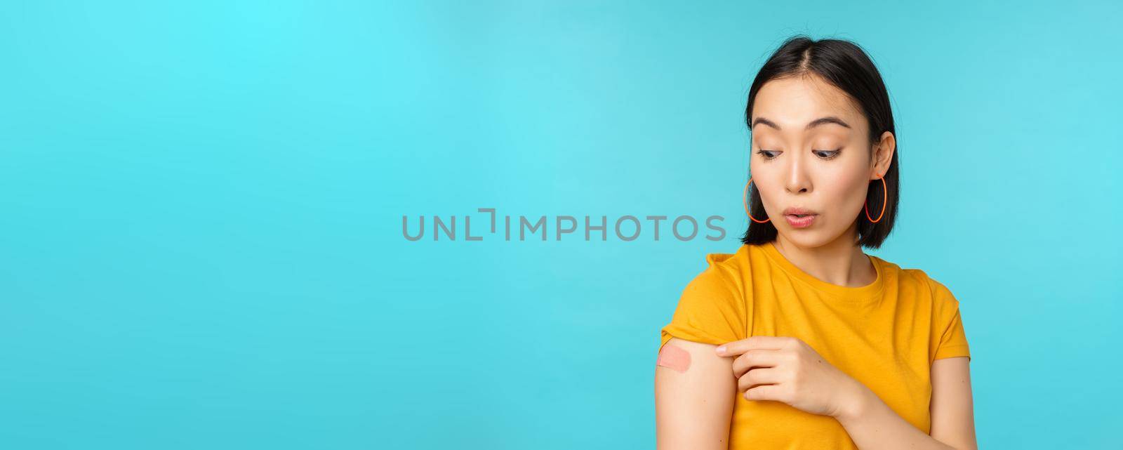 Vaccine campaign from covid-19. Young beautiful, healthy asian woman showing shoulder with bandaid, concept of vaccination, standing over blue background.