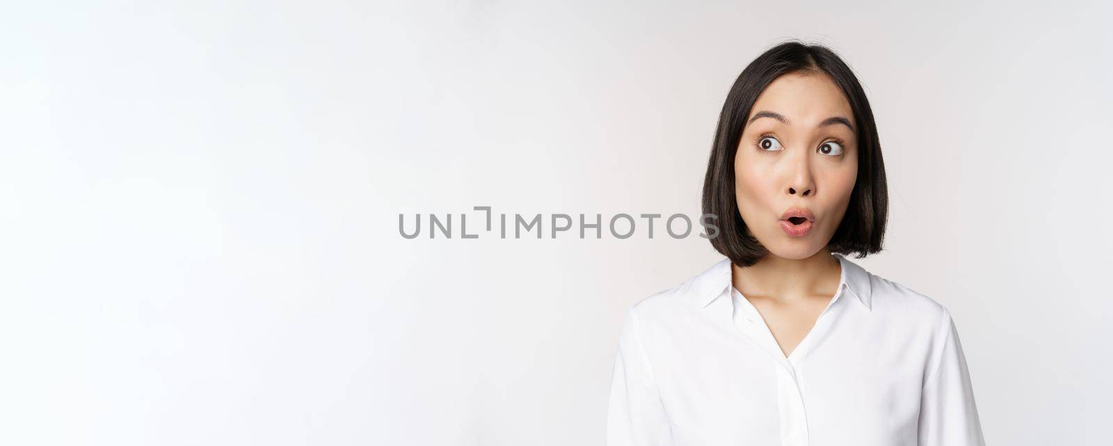 Close up portrait of young asian woman making surprised face, looking at upper left corner impressed, wow emotion, standing over white background by Benzoix