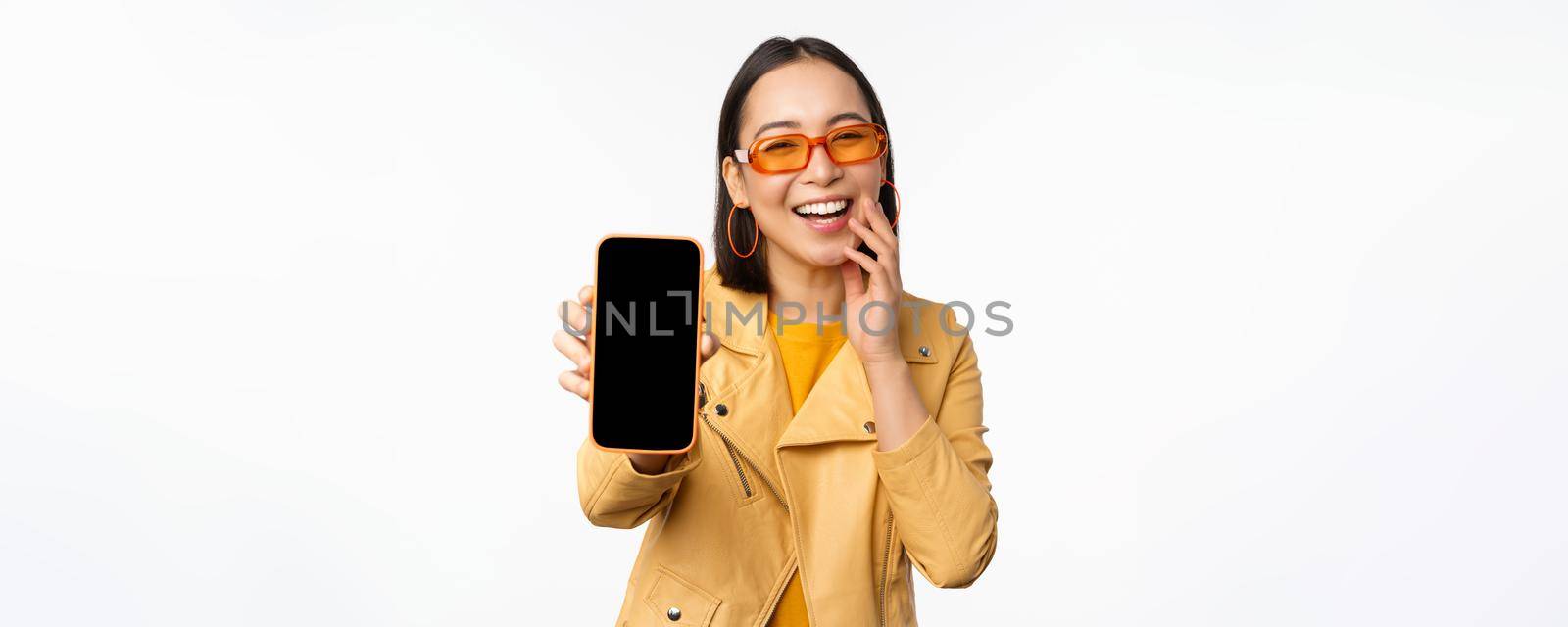 Enthusiastic asian female model, showing smartphone app interface, online store or website on mobile phone screen, standing over white background.