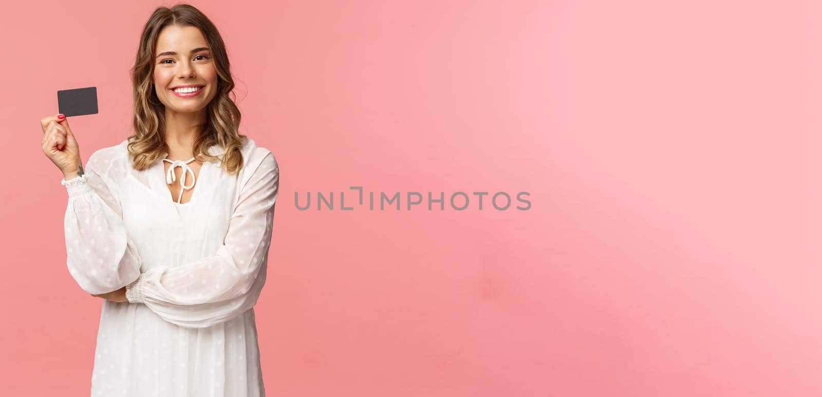 Portrait of pleased good-looking blond european female in white dress, show credit card with satisfied expression, smiling camera, recommend bank services, use payment online, pink background.
