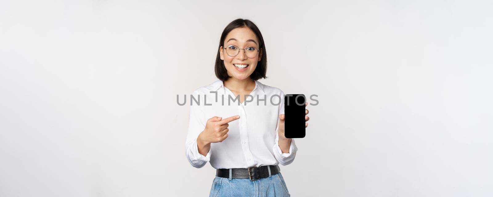 Enthusiastic young asian woman pointing finger at smartphone screen, showing advertisement on mobile phone, white background.