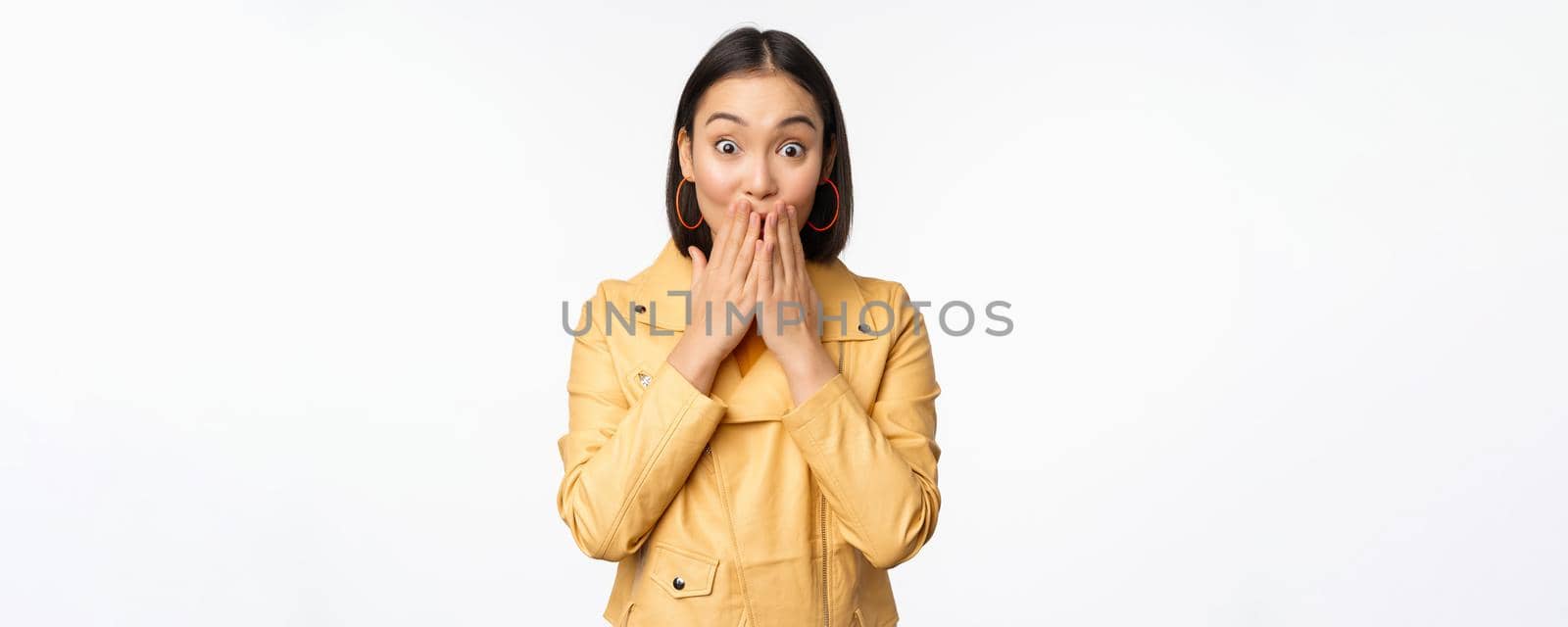 Portrait of excited asian girl looking with interest at camera, amazed reaction at big news or announcement, standing in yellow jacket over white background by Benzoix