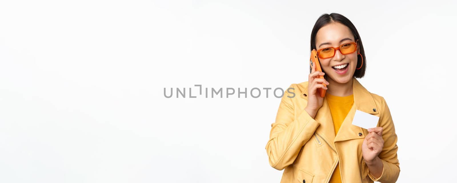 Stylish asian female model, talking on smartphone and showing credit card, standing over white background by Benzoix