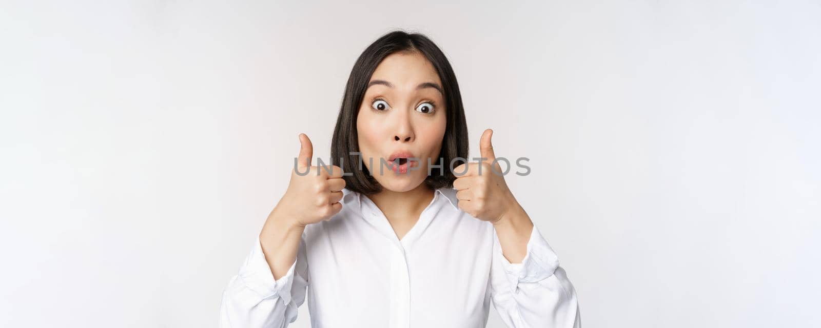 Cheerful asian woman face, showing thumbs up and smiling, pleased by something, recommending smth good, standing over white background by Benzoix