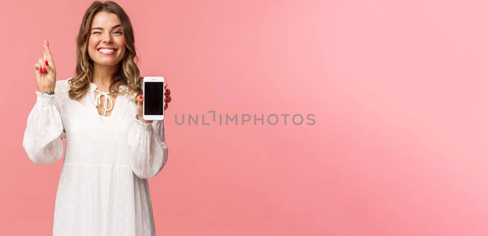 Portrait of lucky and hopeful beautiful blond girl in white dress, making bet, showing mobile phone display, cross finger good luck and smiling optimistic, have faith she will win in competition by Benzoix