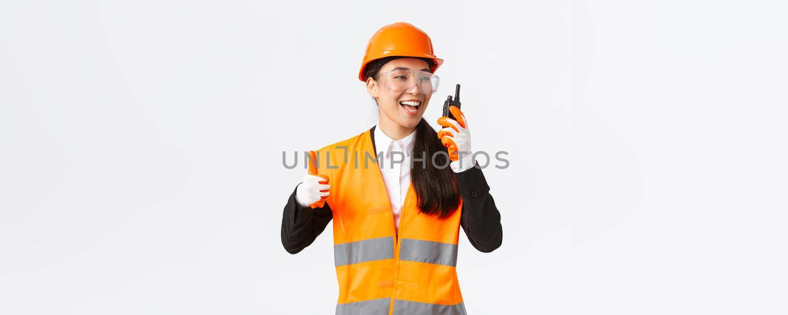 Satisfied happy smiling asian female engineer, industrial technician in safety helmet and uniform showing thumbs-up while praising great work using walkie-talkie, give permission to work by Benzoix