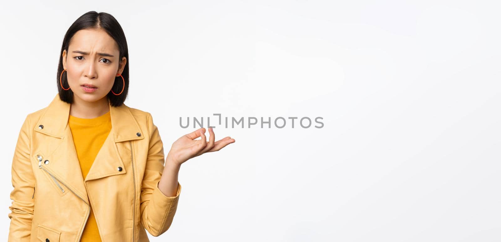 Image of frustrated korean woman, shrugging shoulders and raising hand, looking puzzled, cant understand smth, standing over white background.