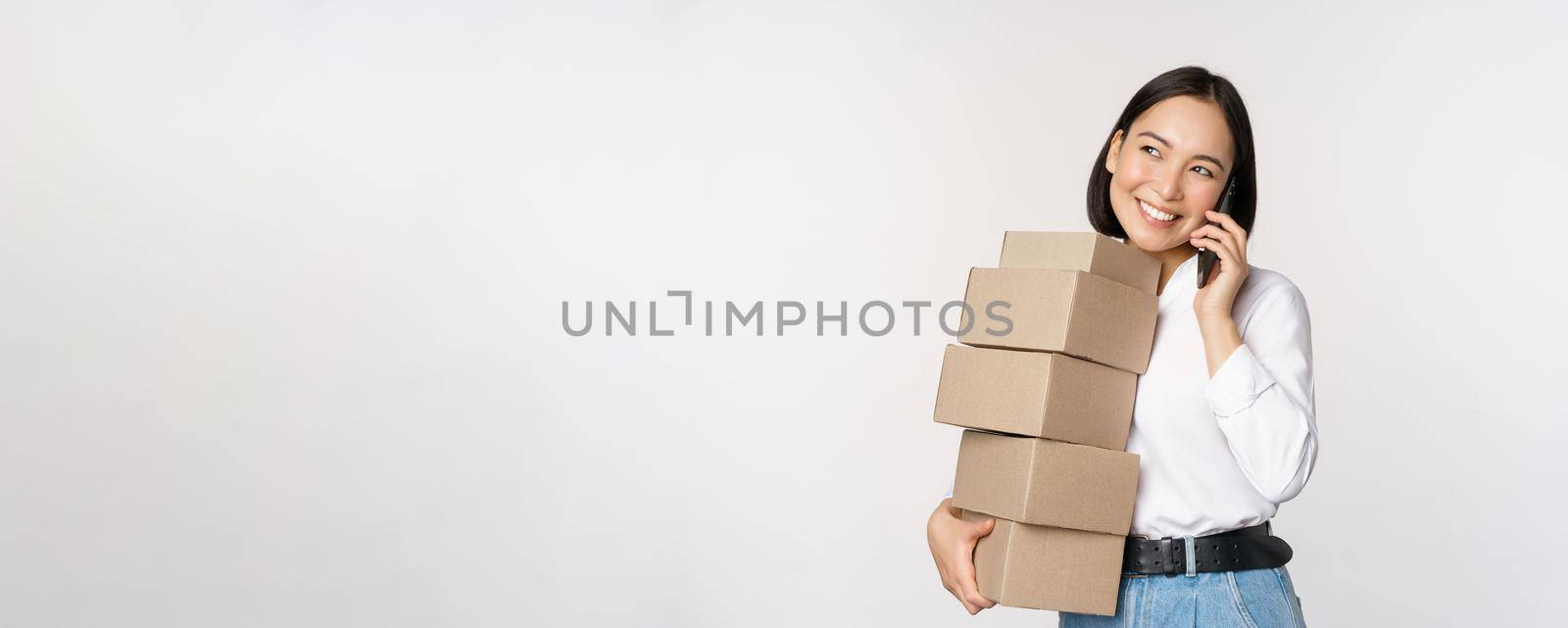 Image of young asian businesswoman answer phone call while carrying boxes for delivery, posing against white background.