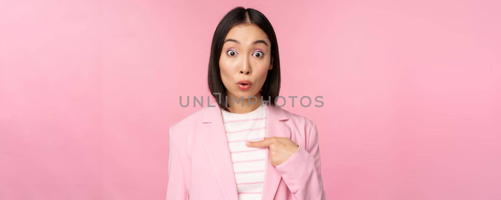 Portrait of asian businesswoman reacts surprised, points at herself with disbelief on face, posing in suit against pink background.