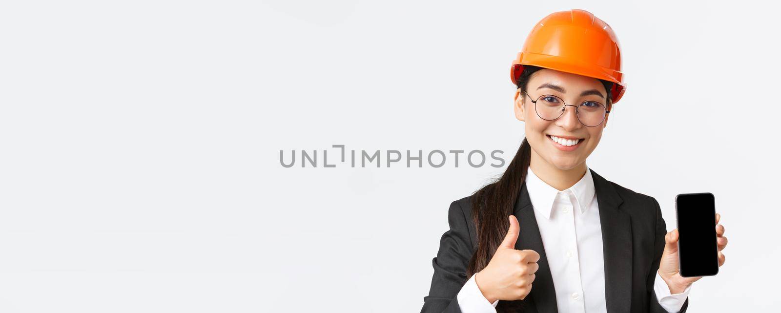 Close-up of professional smiling female engineer, construction manager in business suit and safety helmet, showing smartphone screen and thumbs-up in approval, white background.