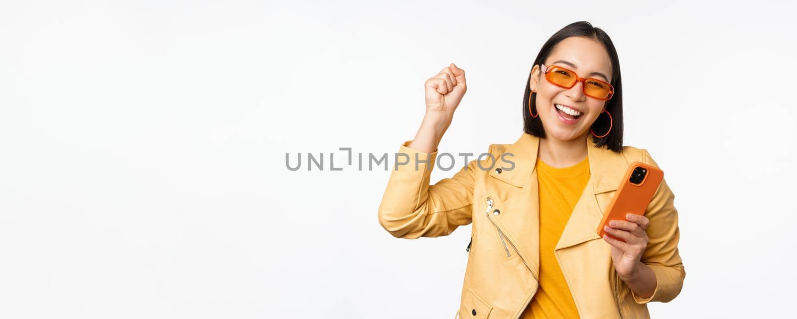 Enthusiastic korean girl in sunglasses, holding smartphone, celebrating and dancing, laughing happy with mobile phone, standing over white background.