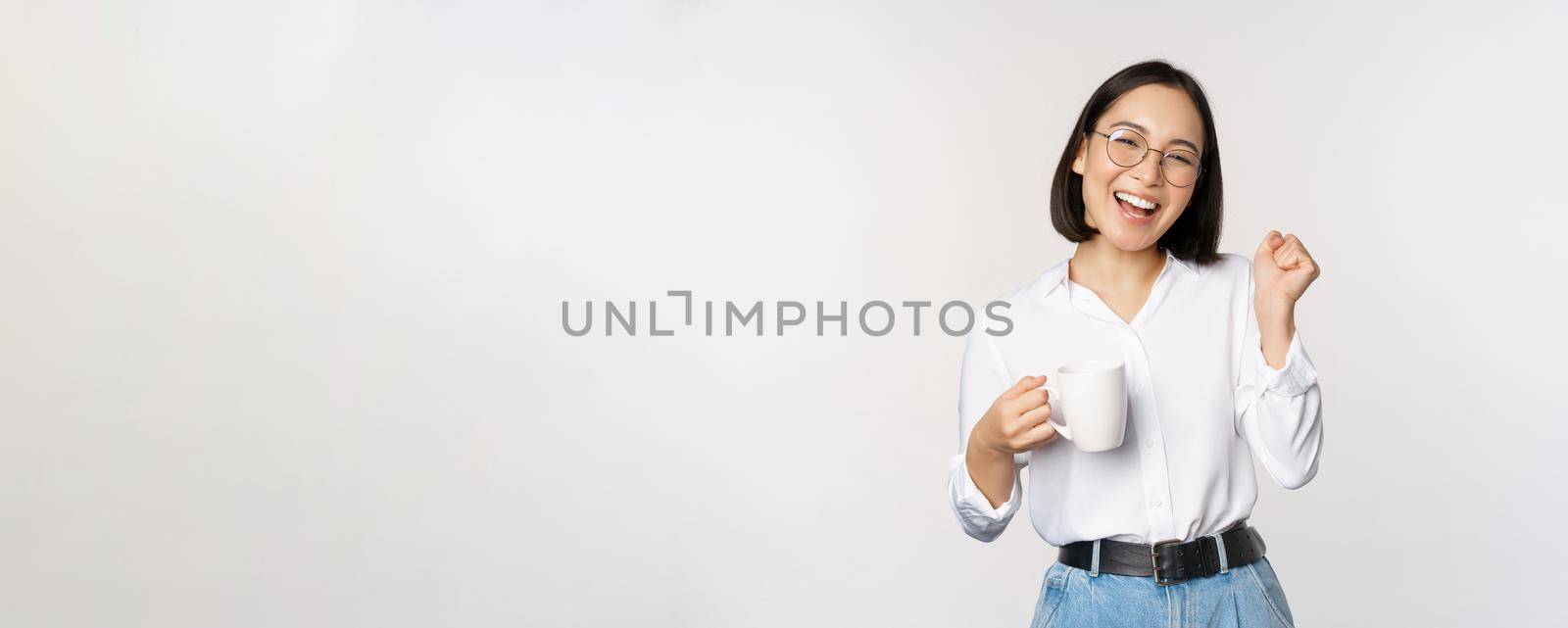 Happy dancing woman drinking coffee or tea from mug. Korean girl with cup, standing over white background by Benzoix