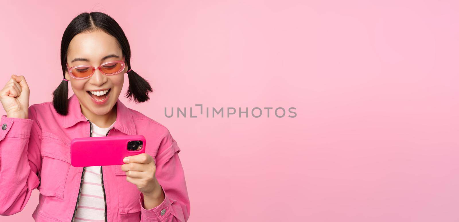Happy smiling korean girl winning on mobile phone, looking at horizontal smartphone screen and rejoicing, achieve goal, celebrating, standing over pink background by Benzoix