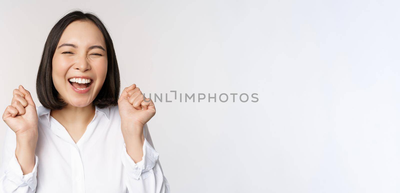 Close up face portrait of dancing asian woman smiling, triumphing and celebrating with happy emotion, standing over white background. Copy space