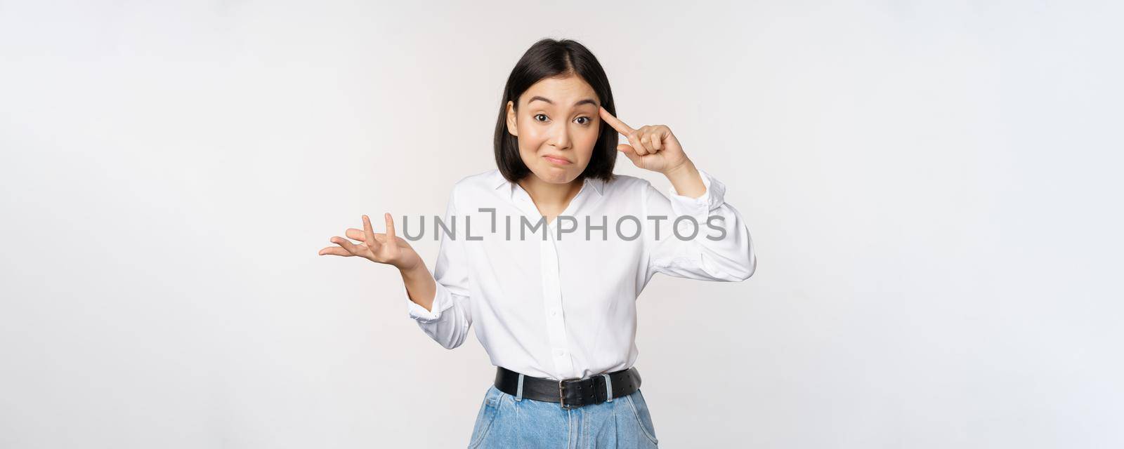 Are you insane. Portrait of frustrated young asian woman, roll finger near head and srugging, someone crazy or stupid, standing over white background.