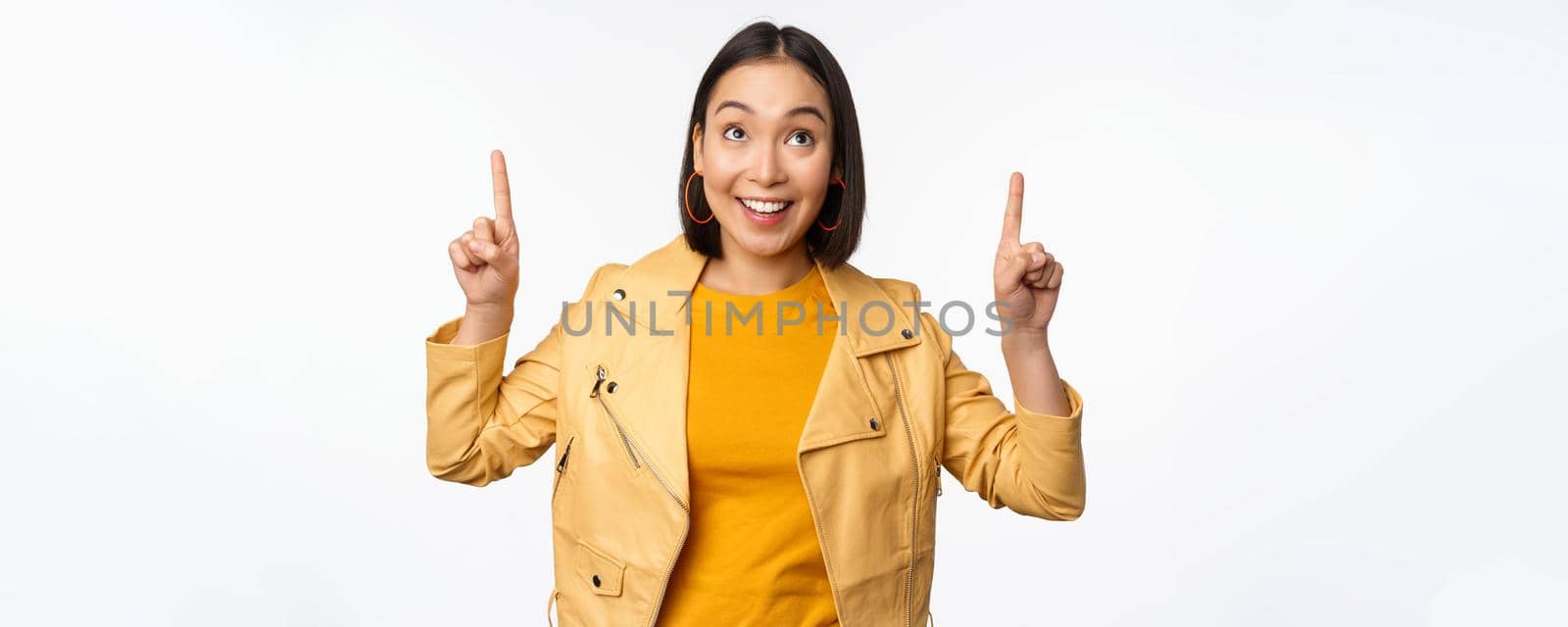 Image of smiling asian brunette woman pointing fingers up, showing advertisement with happy face, posing against white background.