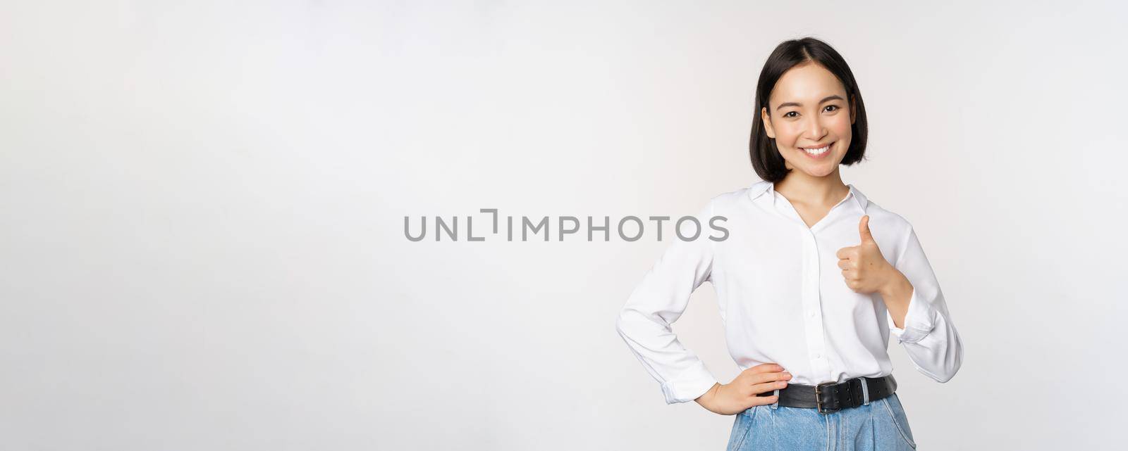 Image of confident asian woman showing thumb up in approval, recommending, like smth good, standing over white background.