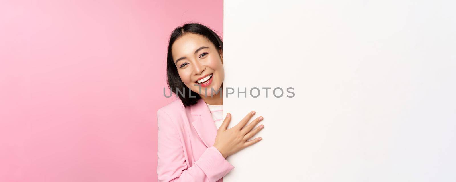 Portrait of young japanese business woman, corporate lady in suit pointing on wall with chart, showing diagram or advertisement on empty copy space, pink background.
