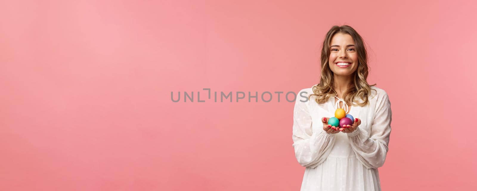 Holidays, spring and party concept. Portrait of tender, lovely blonde girl in white dress, holding Easter painted eggs, celebrating orthodox day, smiling cheerful share positivity, pink background.