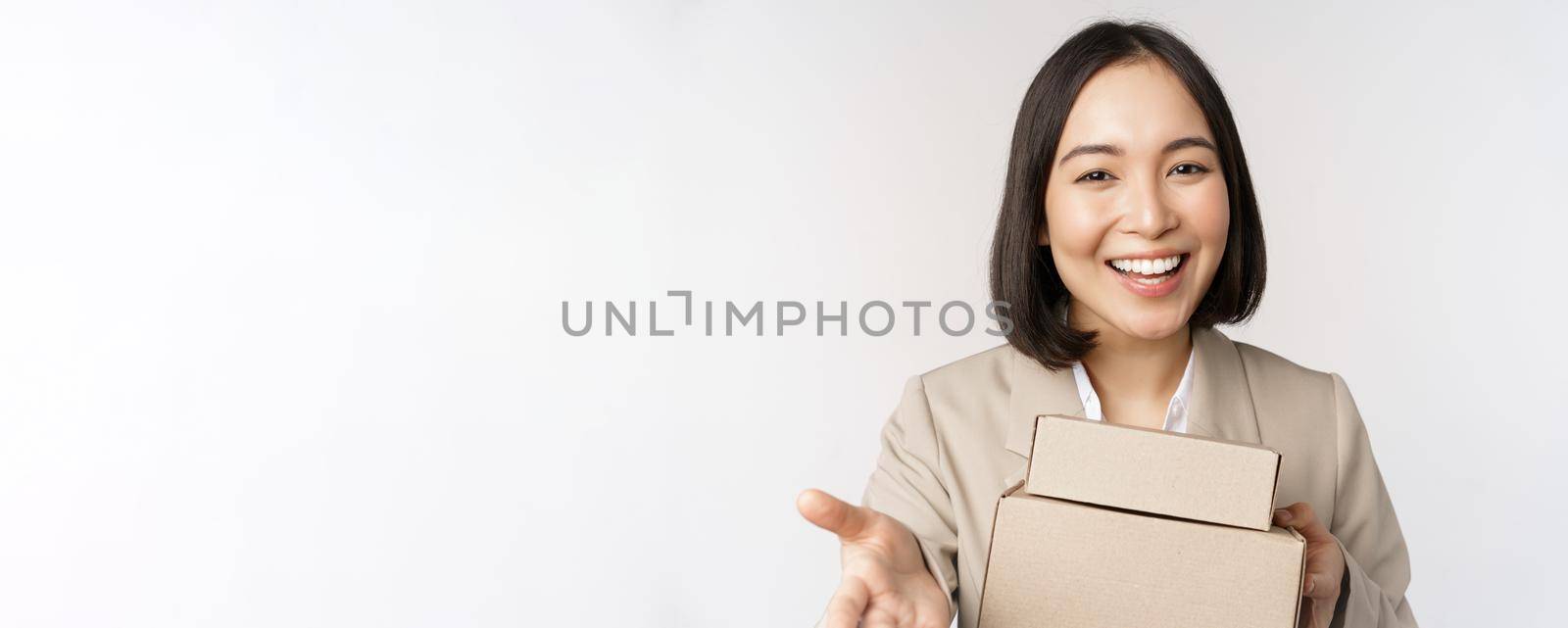 Portrait of business woman, asian saleswoman pointing at you, giving boxes with orders, standing in suit over white background by Benzoix