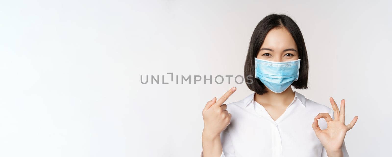 Portrait of asian girl in medical mask showing okay sign and pointing at her covid protection, standing over white background. Copy space