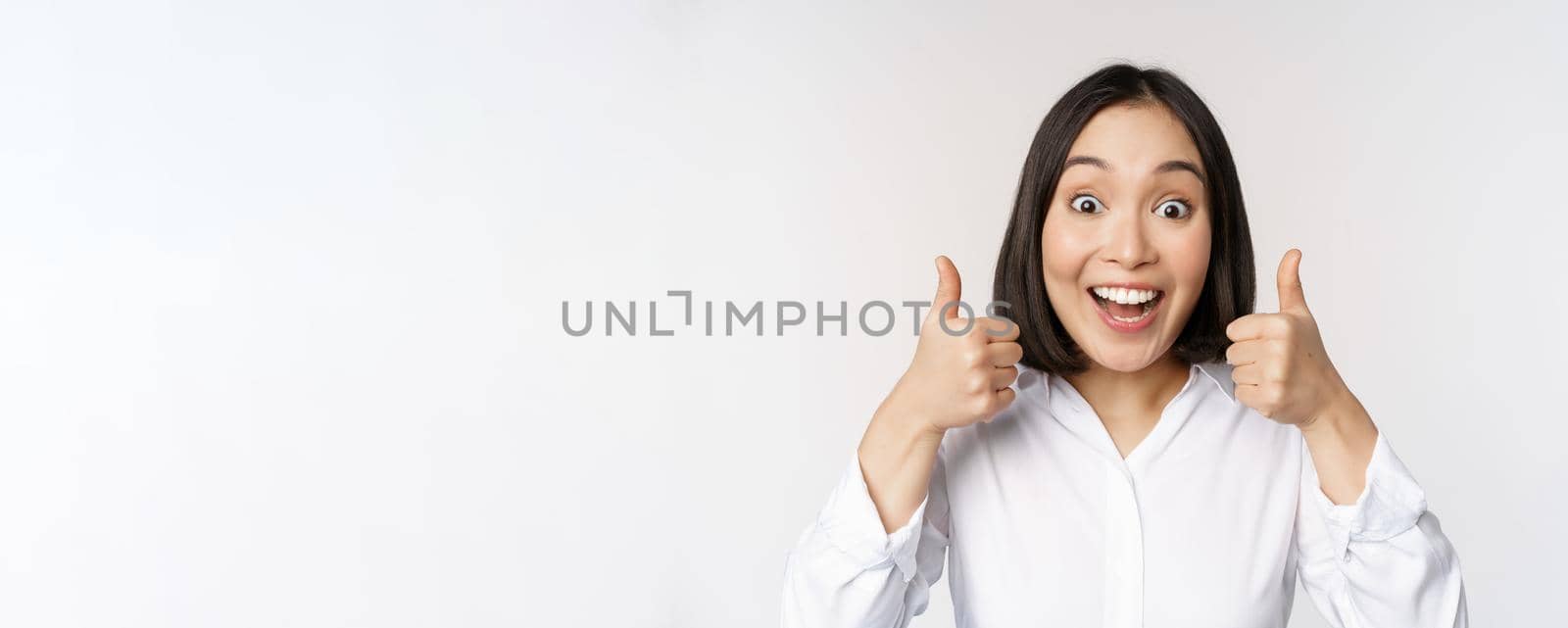 Enthusiastic asian girl showing thumbs up and smiling, pleased by something, recommending smth good, standing over white background.