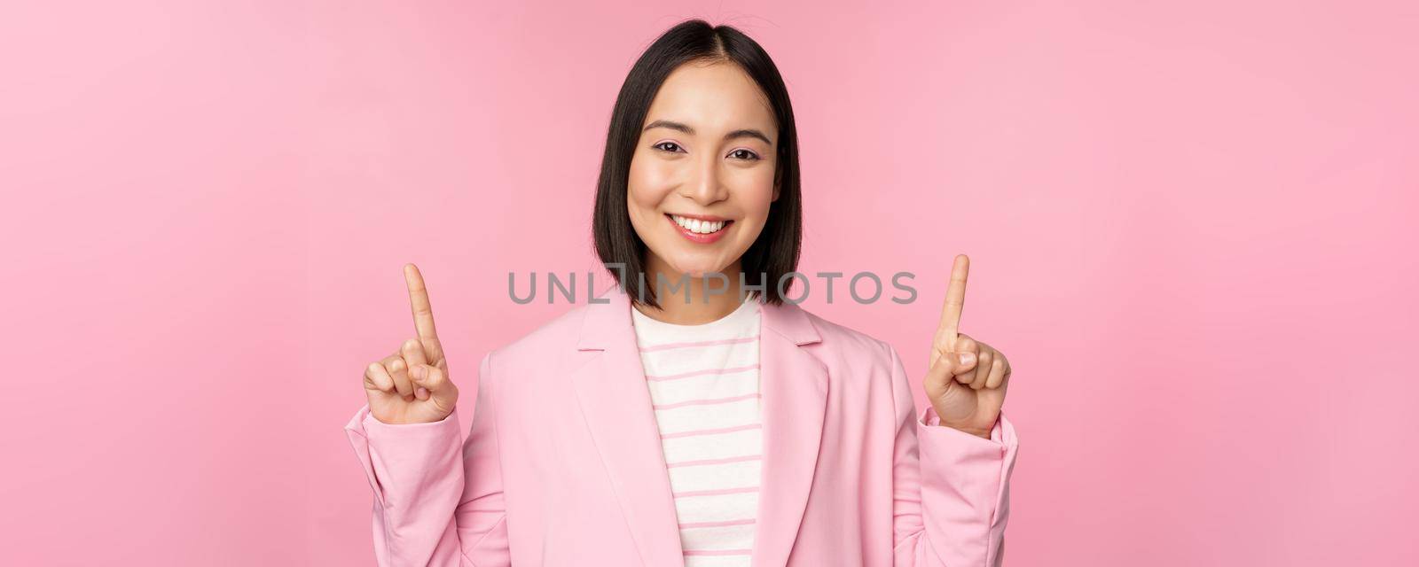 Portrait of asian businesswoman pointing fingers up and smiling, showing business company logo, information on top, standing over pink background by Benzoix