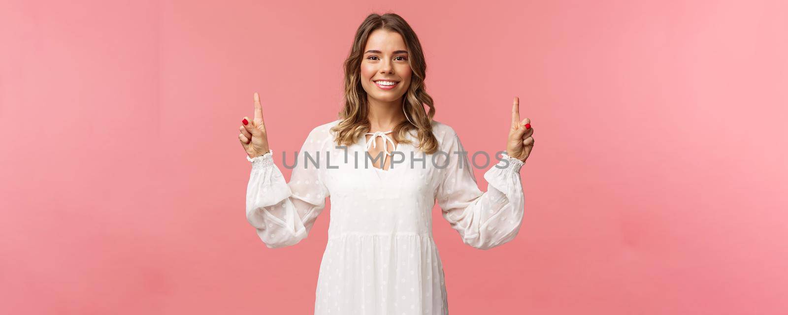 Portrait of confident beautiful young blond woman in white cute dress, pointing fingers up at top advertisement, looking at camera with beaming smile, standing pink background.