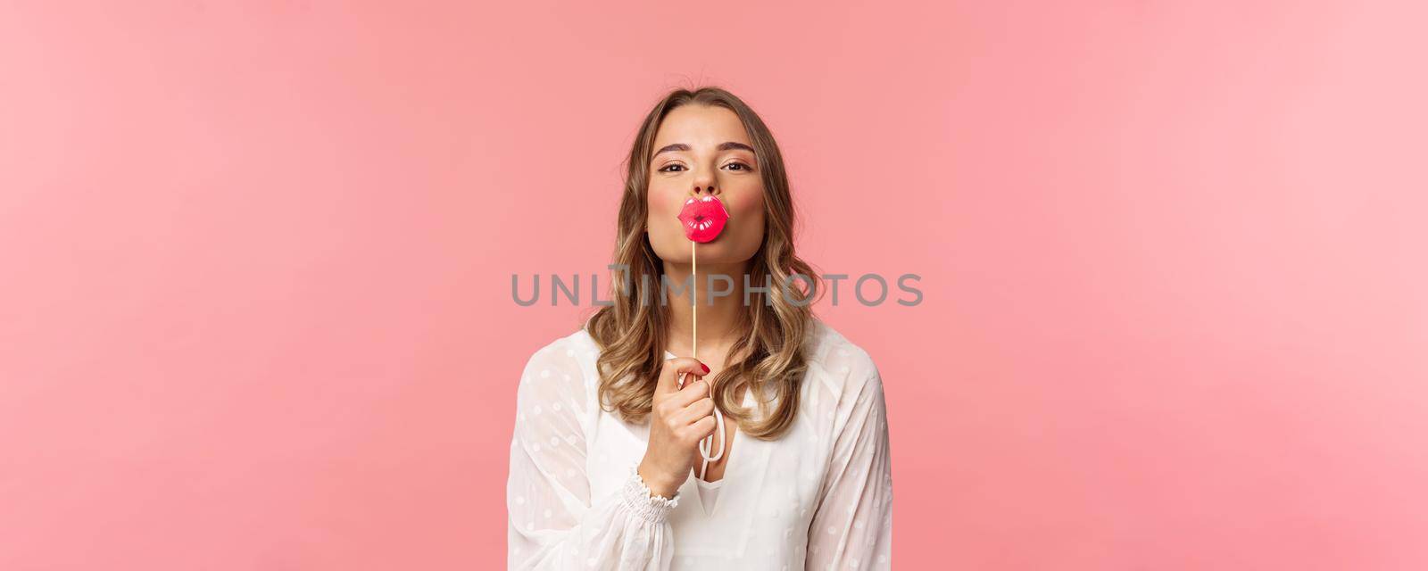 Spring, happiness and celebration concept. Close-up of romantic attractive blond caucasian girl in white dress, holding big kissing lips stick over mouth and looking lovely camera, pink background.