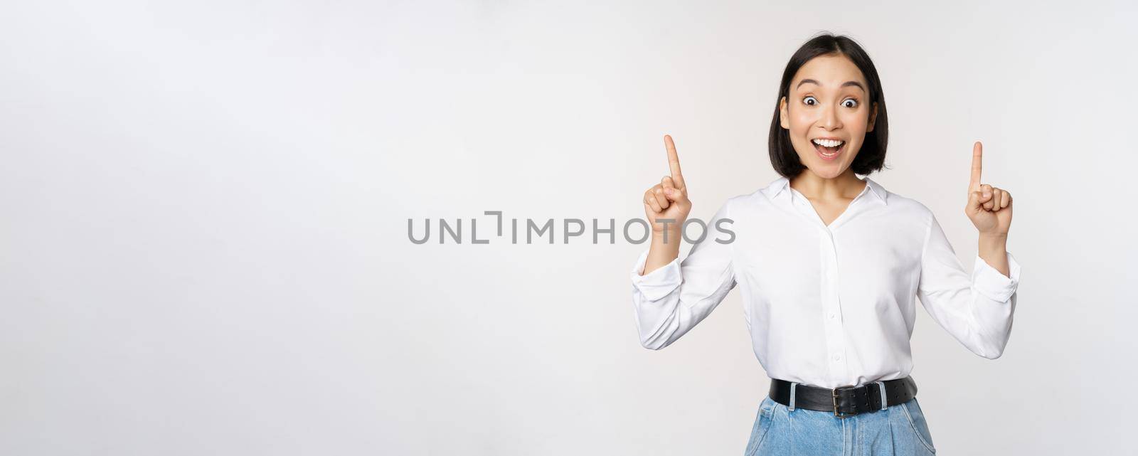 Enthusiastic business woman, asian female model pointing fingers up and smiling, making announcement, showing logo banner on top, white background by Benzoix