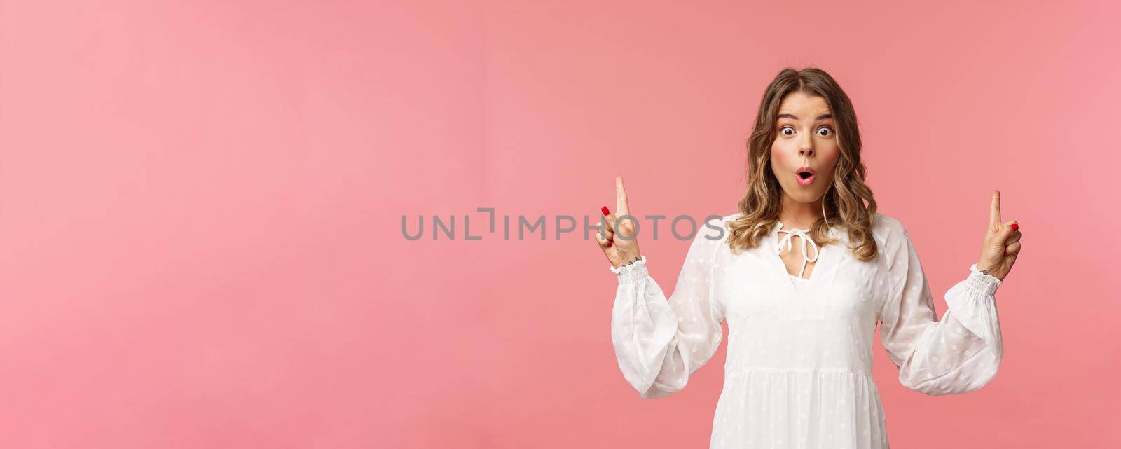 Waist-up portrait of amazed blond girl say wow in excitement, pointing fingers up, showing person top advertisement with cool special spring discount, standing pink background.