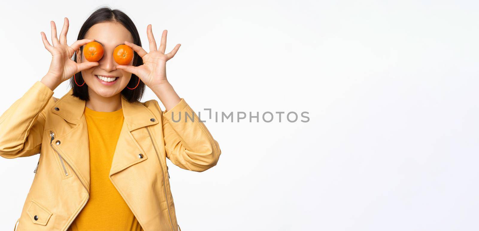 Funny asian girl holding tangerine on eyes and smiling, making playful grimaces, standing over white background. Copy space