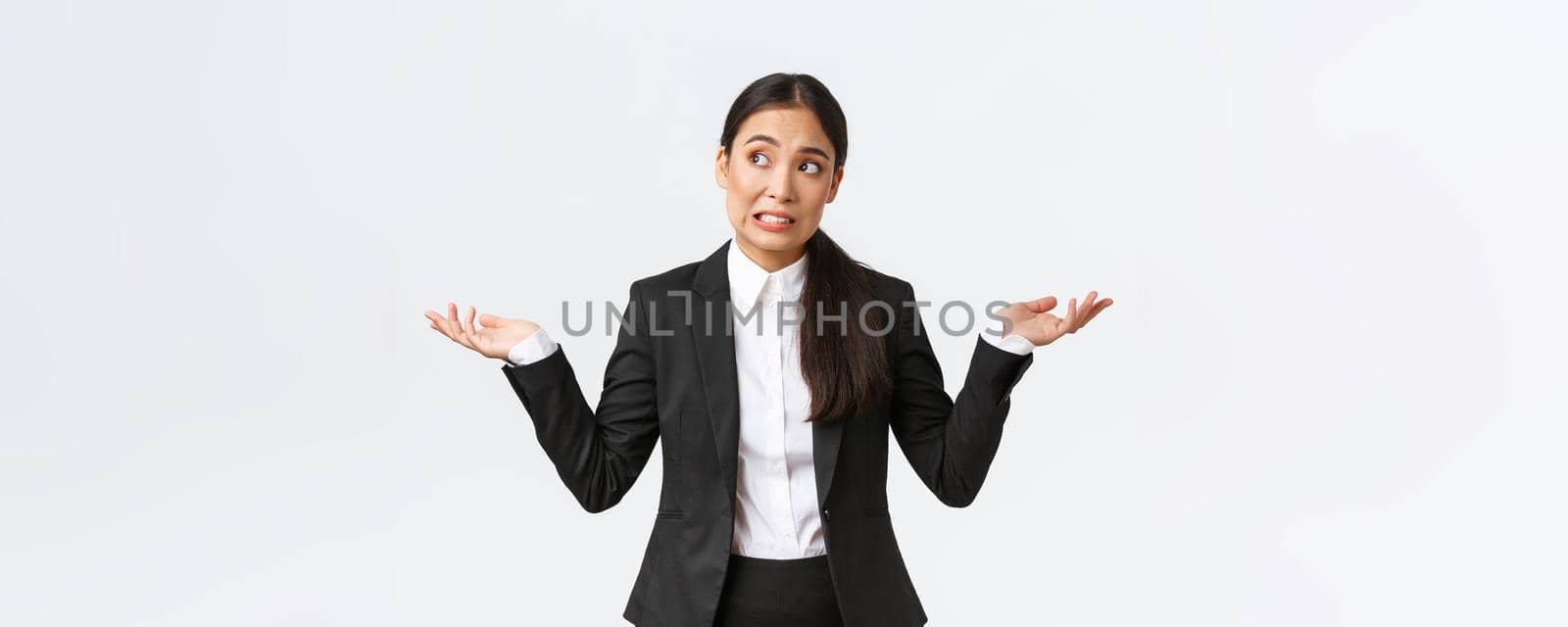 Troubled young female office assistant, saleswoman in black suit shrugging and looking away worried, dont know what do, being confused and unaware, standing nervous over white background by Benzoix