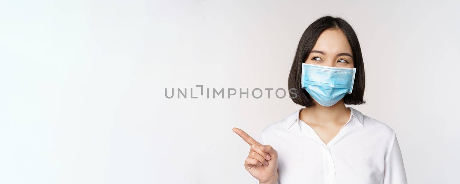 Portrait of cute asian woman in medical face mask, coronavirus protection, pointing finger left and looking intrigued at empty copy space, white background.