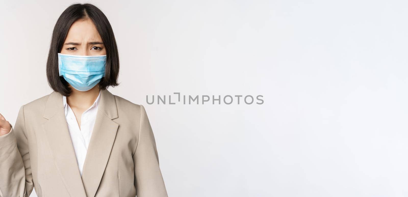 Coronavirus and work concept. Frustrated asian business woman in medical face mask, pointing finger left and complaining, standing over white background by Benzoix