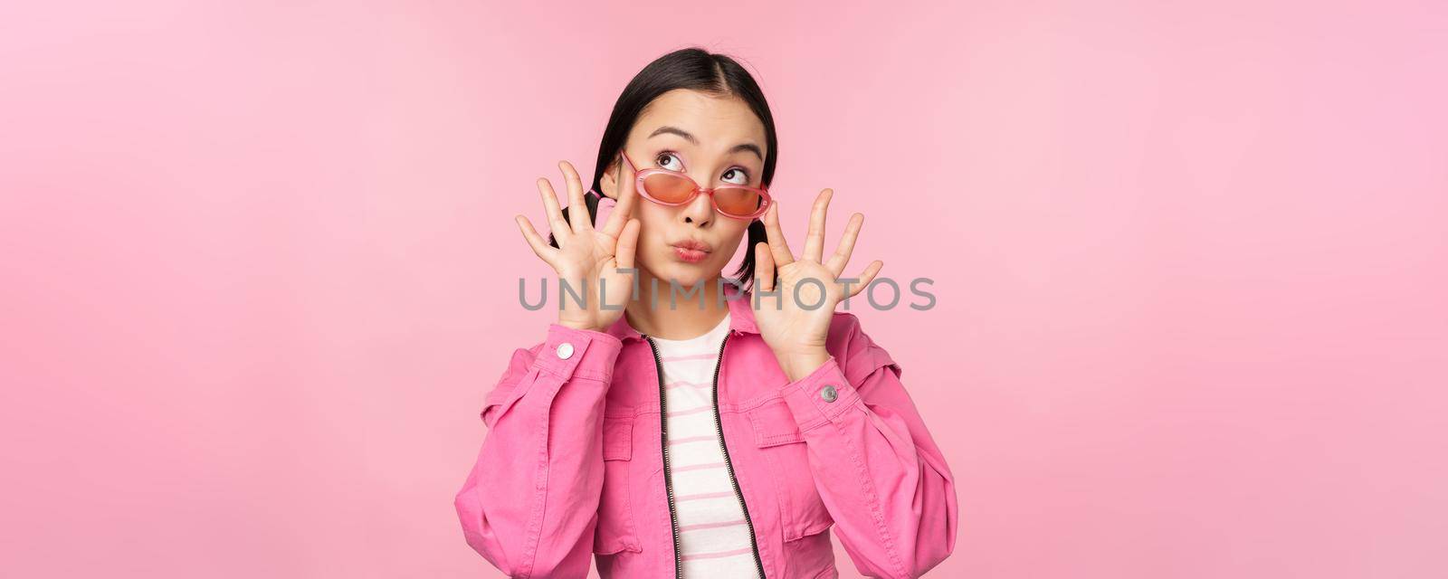 Close up of beautiful asian female model in stylish sunglasses, posing against pink background in trendy outfit, copy space.