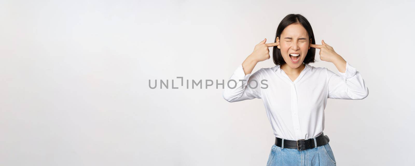 Annoyed asian woman feeling discomfort from loud annoying noise, close ears and eyes, standing over white background by Benzoix