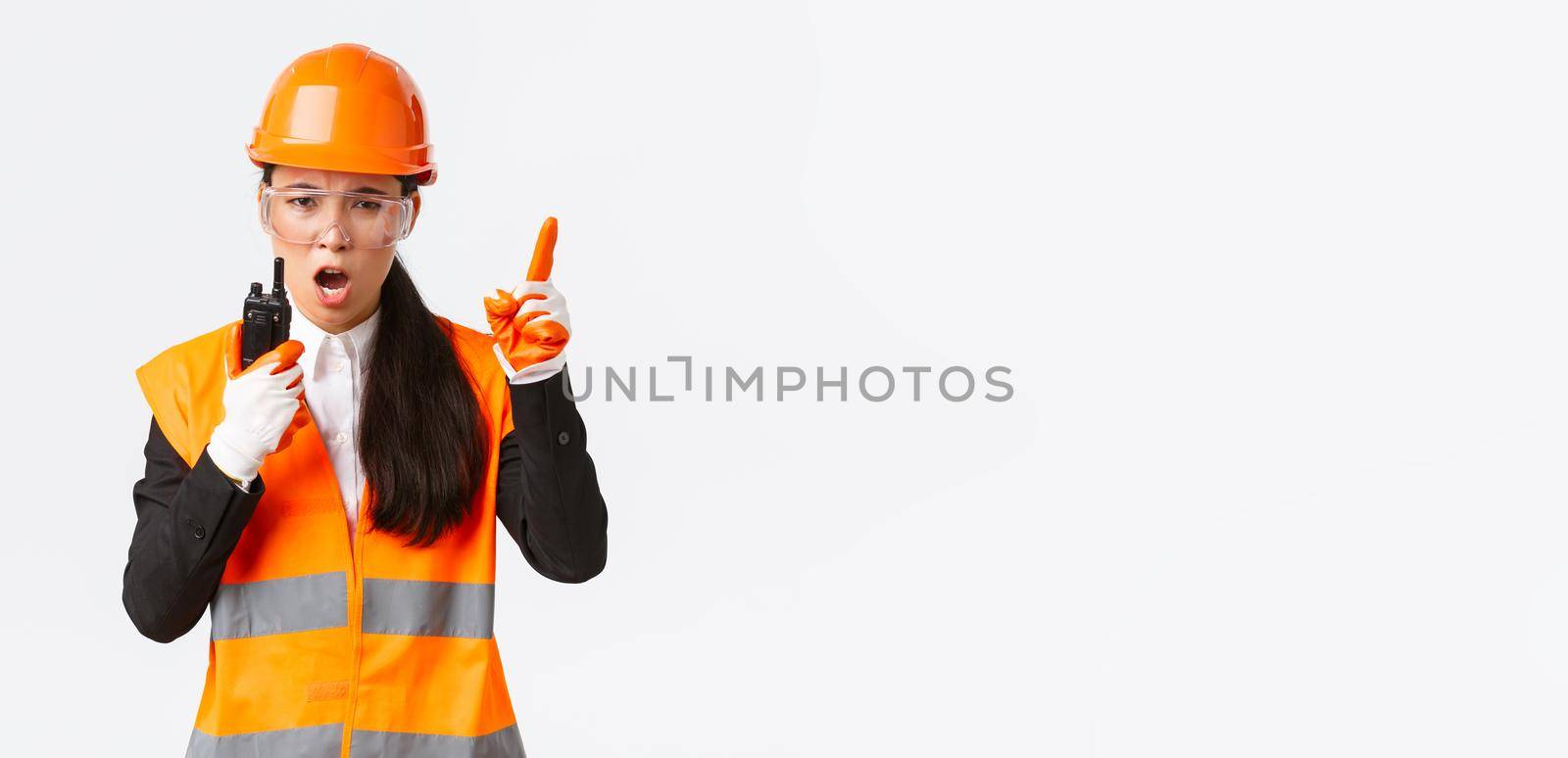Serious-looking asian female industrial engineer, technician in safety helmet and uniform command construction team with walkie-talkie, explain or scolding someone with radio phone.