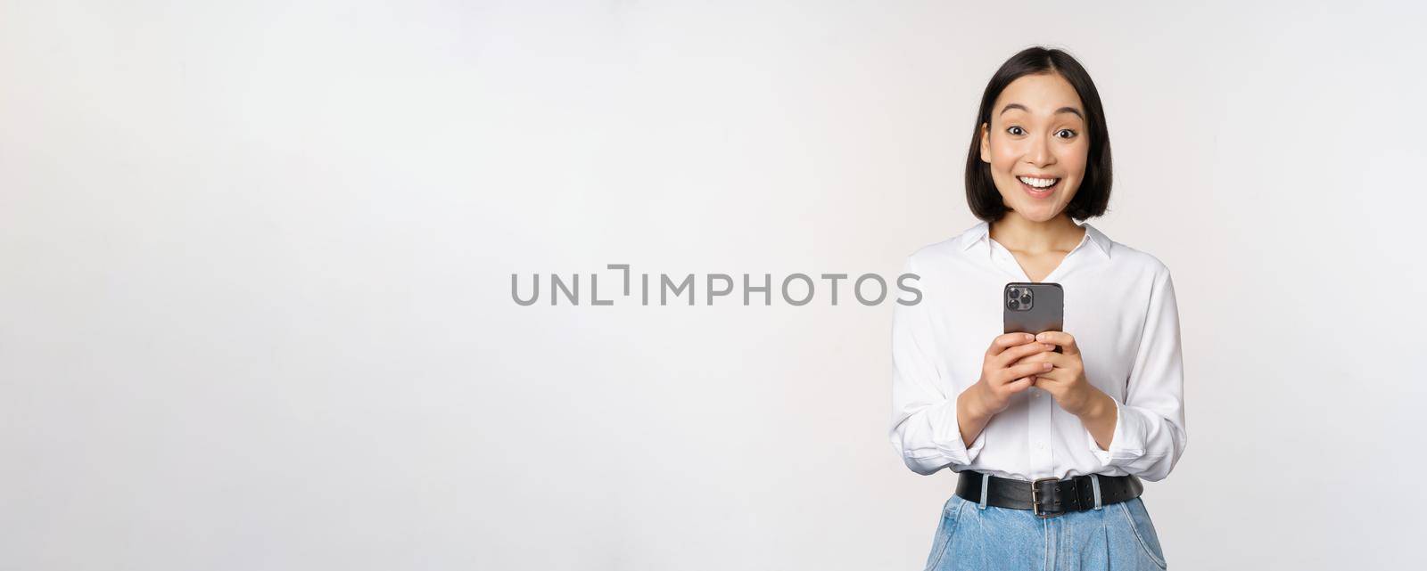 Excited asian woman smiling, reacting to info on mobile phone, holding smartphone and looking happy at camera, standing over white background.