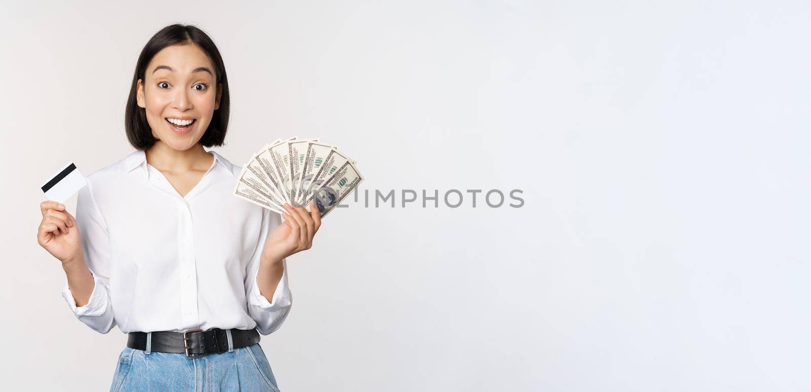 Portrait of enthusiastic asian woman holding money in cash and credit card, smiling amazed at camera, white background by Benzoix