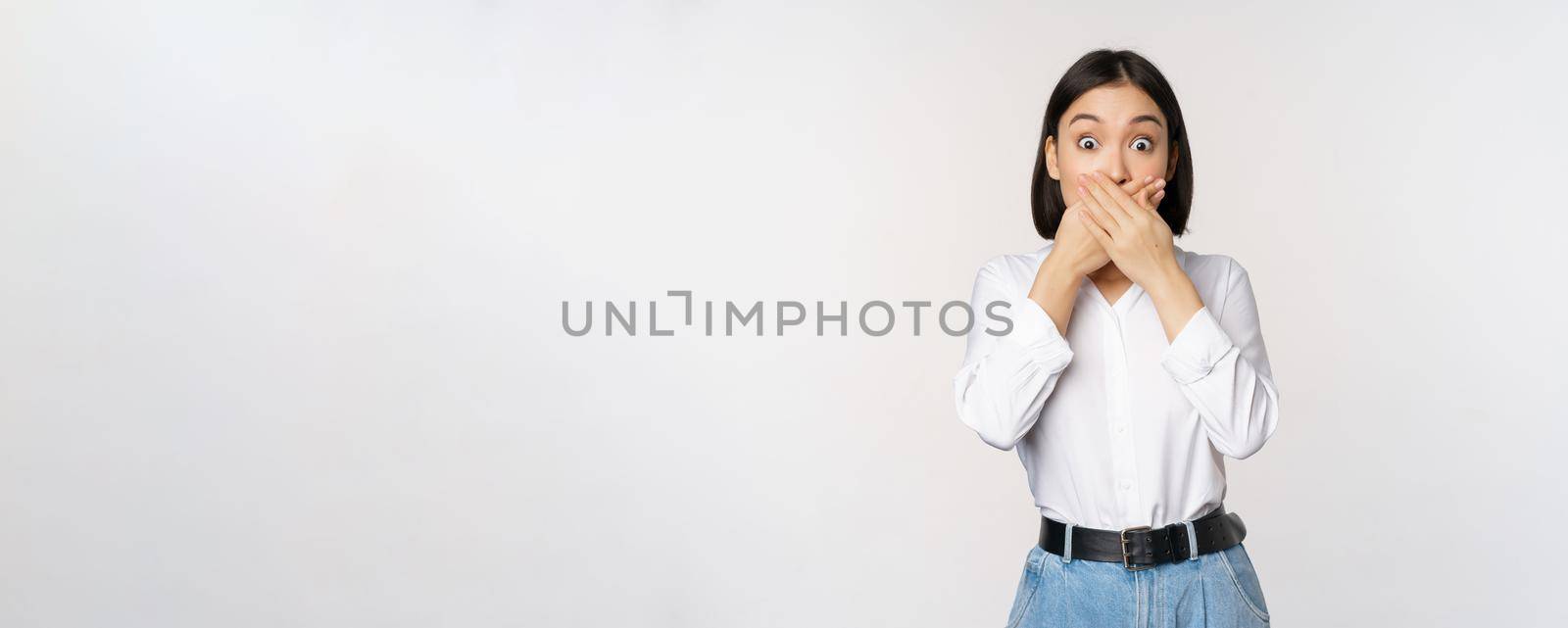 Shocked young asian girl close mouth, cover lips with hands and looking amazed, standing over white background. Taboo gesture