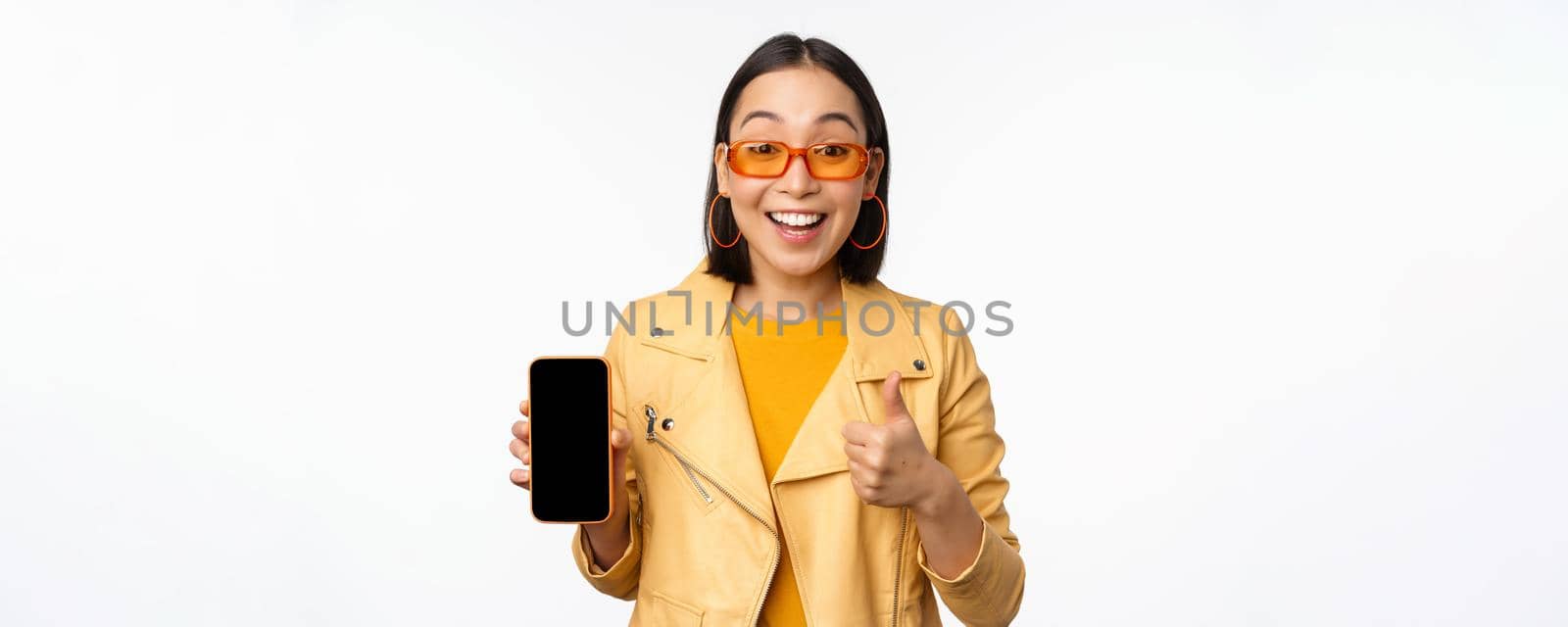 Beautiful korean girl, asian woman in sunglasses, showing smartphone app interface, thumbs up, recommending mobile phone application, white background.