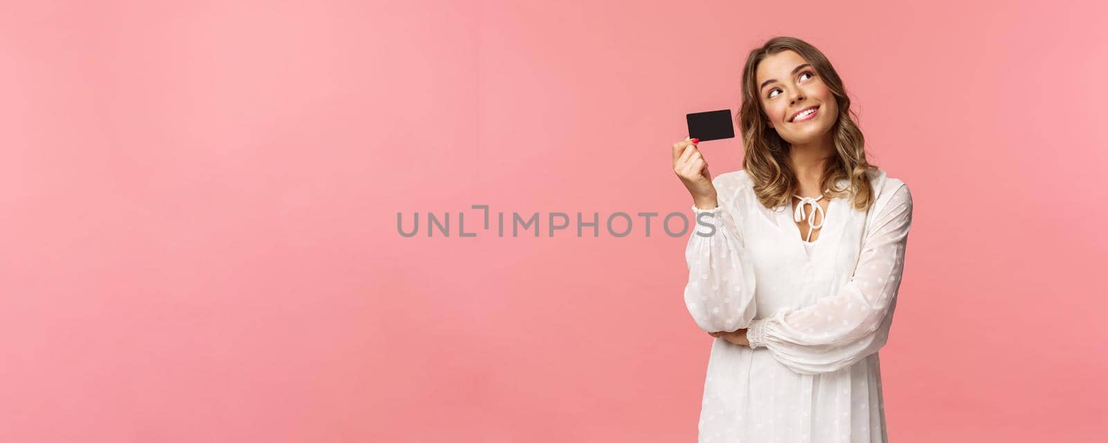 Portrait of cute tender feminine blond girl daydreaming about how she will waste her money on online shopping, look up dreamy, imaging as waiting delivery, hold credit card, pink background.