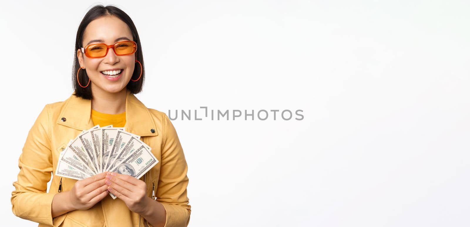 Stylish smiling asian girl holding money cash, showing dollars and celebrating, standing over white background. Copy space