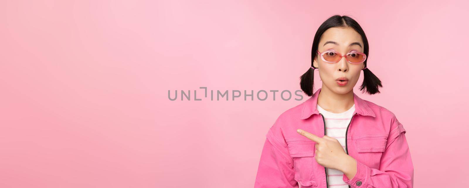 Close up portrait of asian woman gasping, looking surprised, pointing finger at banner, advertisement, standing over pink background.