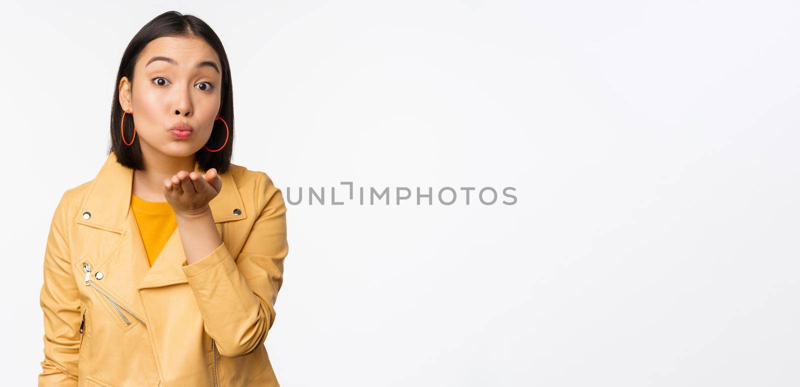 Cute korean stylish girl, sending air kiss, blowing mwah and looking silly romantic at camera, standing over white background. Copy space