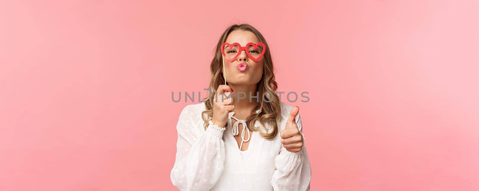 Spring, happiness and celebration concept. Close-up portrait sassy and coquettish attractive blond girl in white dress, holding heart-shaped glasses on eyes, folding lips in kiss and show thumbs-up.