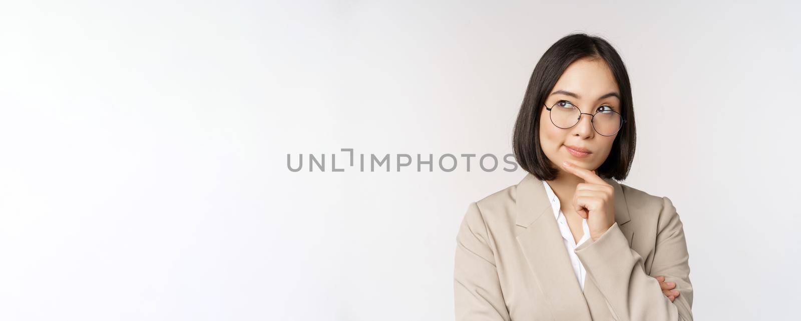 Portrait of thoughtful asian businesswoman in glasses, making assumption, thinking, standing in beige suit against white background.