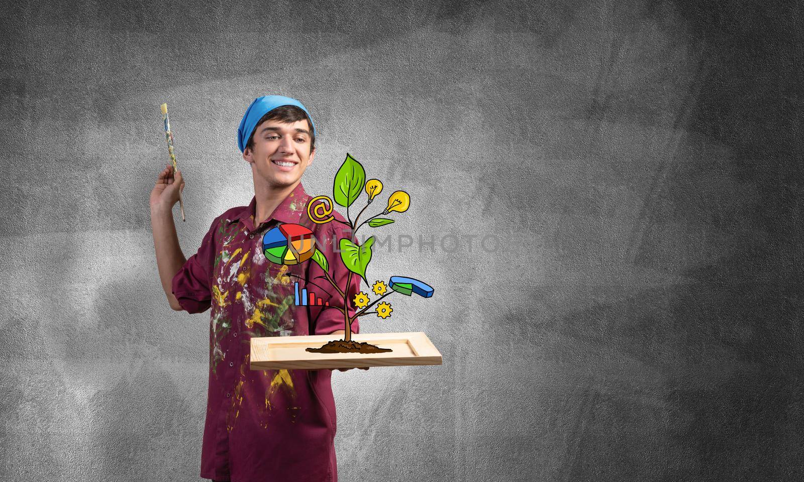 Young artist drawing plant of wealth and business success. Happy painter in dirty shirt and bandana standing on grey wall background. Original business idea generation and development.