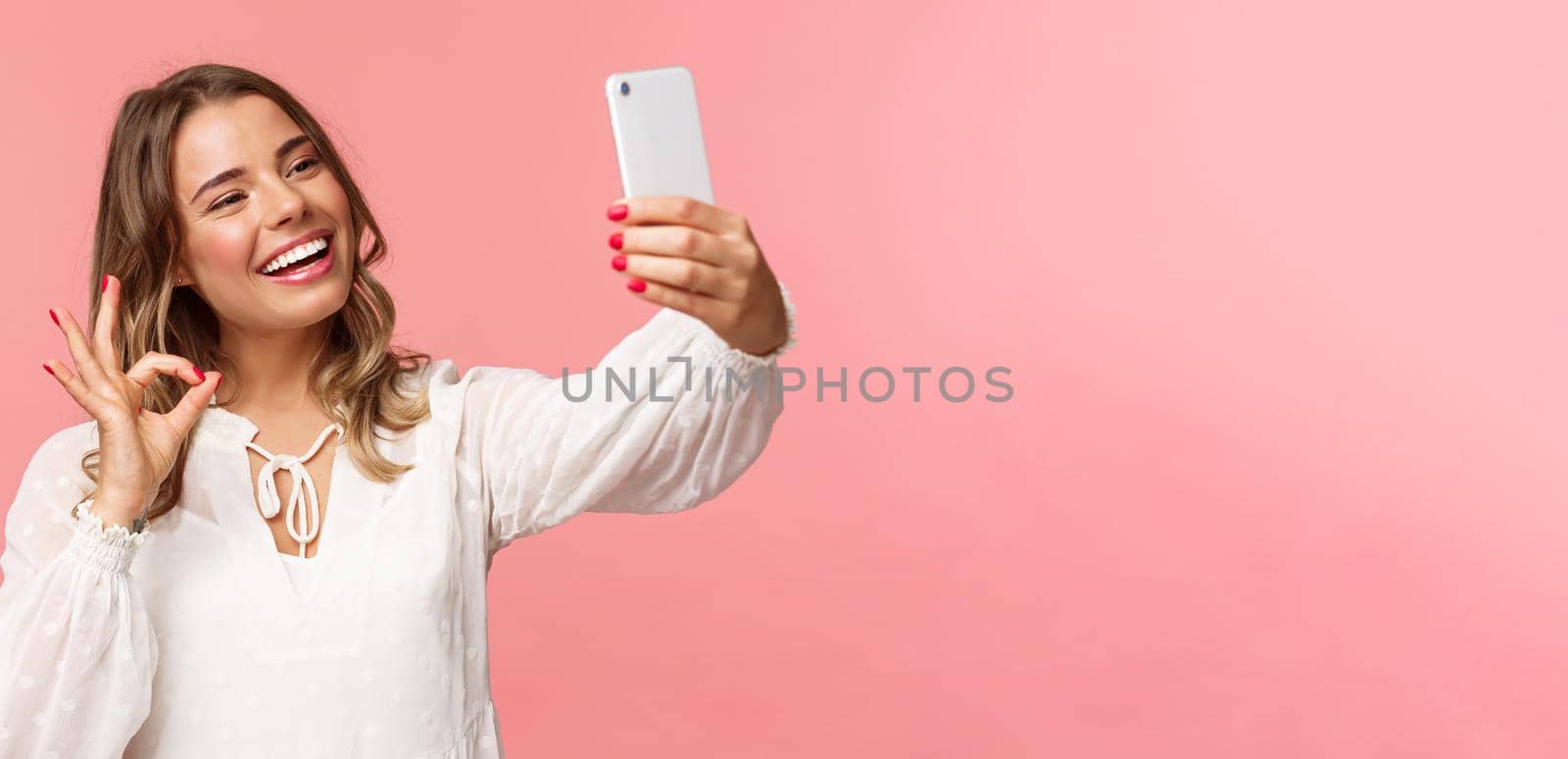 Close-up of satisfied good-looking blond girl in white dress, taking selfie, record mobile phone video, show okay satisfactory sign with pleased nod, smiling agree or recommend, pink background.